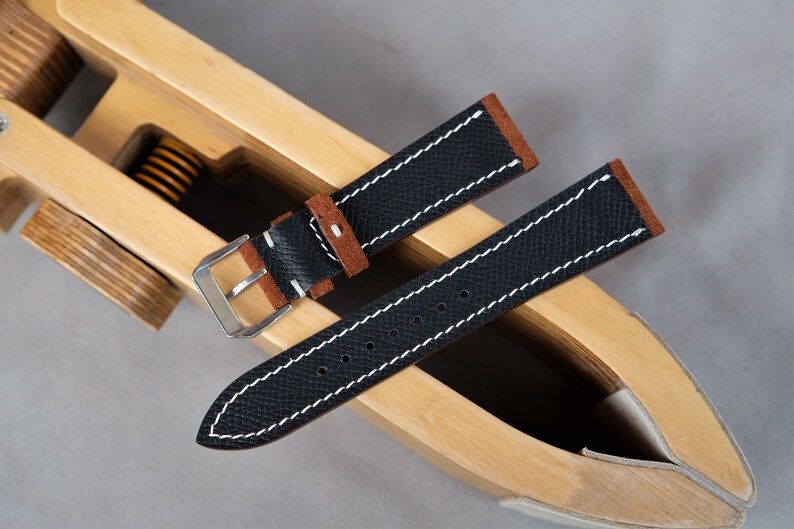 Puede incluir: Dos correas de reloj de cuero negro con costuras blancas. La correa de arriba tiene un forro de ante marr&oacute;n. Las correas est&aacute;n en una herramienta de correa de reloj de madera.