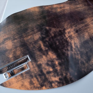 Könnte beinhalten: Ein dunkelbraunes Lederarmband mit abgenutzter Oberfläche. Das Armband liegt auf einem Stück braunem Leder mit abgenutzter Oberfläche.