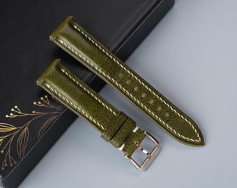 Puede incluir: Dos correas de reloj de cuero verde con costuras blancas. Las correas están sobre una caja negra con diseños de hojas de oro.