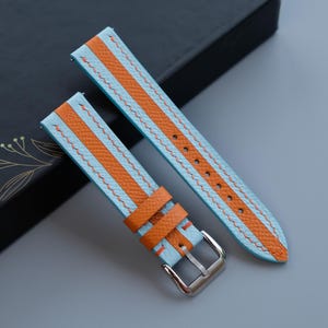 Puede incluir: Correas de reloj con un diseño de rayas azul claro y naranja. Las correas tienen un aspecto texturizado y están hechas de un material duradero. Las correas de reloj tienen una hebilla plateada y se muestran sobre una superficie gris.
