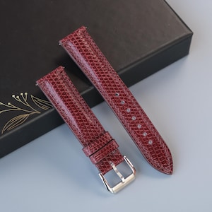 May include: Burgundy watch straps with a textured, reptile-like pattern. The straps are displayed on a black box with gold floral accents. The buckle is silver. The watch straps are new.