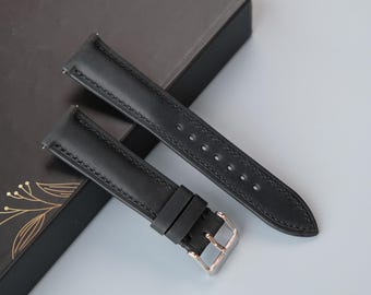 May include: Two black leather watch straps with silver buckles. The straps are lying on a black box with a gold floral pattern.
