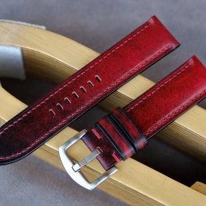 May include: Red leather watch strap with black stitching and a silver buckle. The strap is distressed and has a vintage look.
