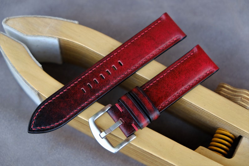 May include: Red leather watch strap with black stitching and a silver buckle. The strap is distressed and has a vintage look.