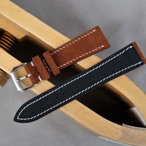Puede incluir: Dos correas de reloj, una de ante marr&oacute;n y otra de cuero negro, con costuras blancas, mostradas en una herramienta de madera para correas de reloj.