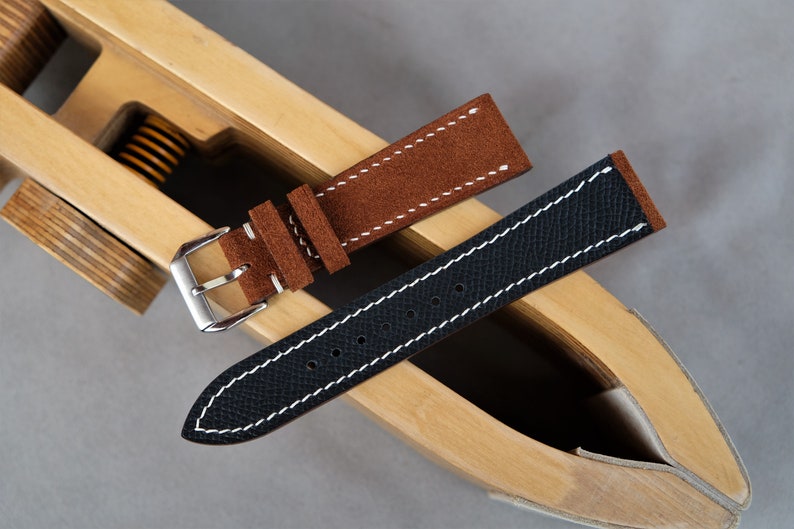 Puede incluir: Dos correas de reloj, una de ante marr&oacute;n y otra de cuero negro, con costuras blancas, mostradas en una herramienta de madera para correas de reloj.