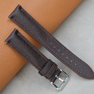 May include: Two dark brown watch straps with reddish-brown stitching and a silver buckle. The straps are made of a textured material and are displayed on a light brown and gray surface. The watch straps are new.