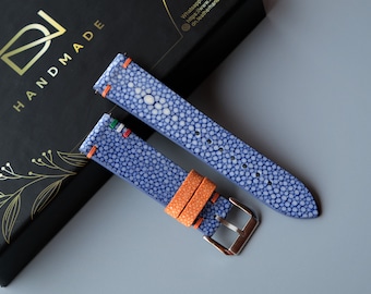 May include: Two blue and white stingray leather watch straps with orange stitching. The straps are on a gray surface. The strap on the left has a small Italian flag on it. The strap on the right has the text "HANDMADE" on a black box.