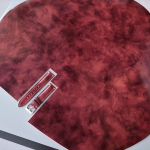 May include: Two burgundy leather watch straps on a white surface. The straps are cut out of a larger piece of leather.