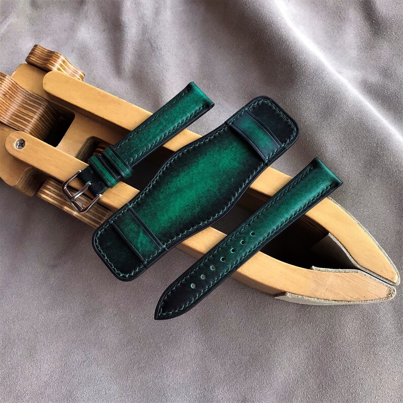 May include: Three green leather watch straps with contrasting black stitching. The straps are laid out on a wooden surface.