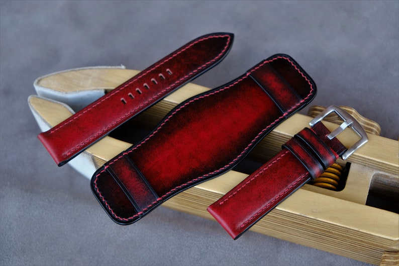 May include: Three red leather watch straps with black stitching. The straps are laid out on a wooden surface. The straps have a distressed look and are made of high-quality leather.