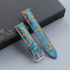 Pode incluir: Duas pulseiras de relógio com um padrão floral azul e laranja. As pulseiras são feitas de um material texturizado e têm fivelas prateadas. São exibidas em uma superfície cinza ao lado de uma caixa preta com detalhes em folhas de ouro.