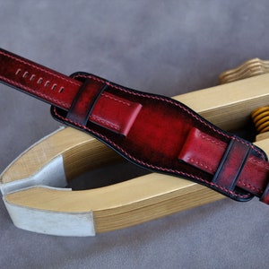 May include: A red leather watch strap with black stitching and a silver buckle. The strap is on a wooden tool.