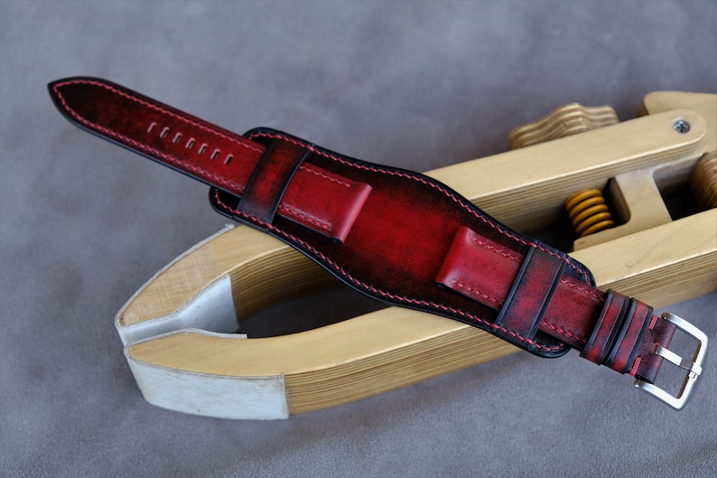 May include: A red leather watch strap with black stitching and a silver buckle. The strap is on a wooden tool.