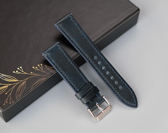 May include: Two dark blue leather watch straps with silver buckles. The straps are lying on a black box with a gold floral design.
