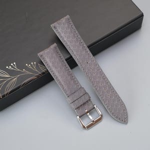 May include: A grey watch strap featuring a repeating hexagonal honeycomb pattern across its surface. The strap includes a silver-toned metal buckle and a matching grey leather keeper loop, designed for attachment to a timepiece case.