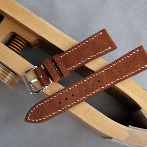 Puede incluir: Dos correas de reloj de cuero marr&oacute;n con costuras blancas. Las correas est&aacute;n dispuestas sobre una herramienta de correa de reloj de madera.