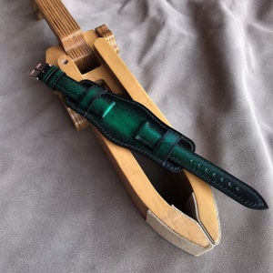 May include: A dark green leather watch strap with black stitching. The strap is on a wooden watch strap forming tool.