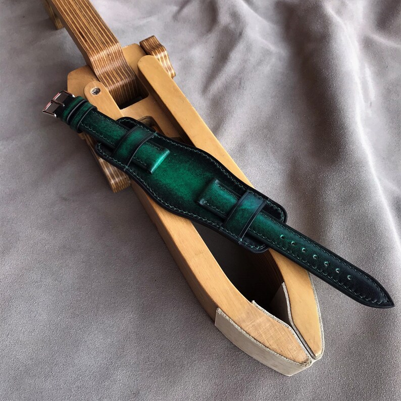 May include: A dark green leather watch strap with black stitching. The strap is on a wooden watch strap forming tool.