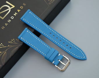 May include: Two light blue leather watch straps with white stitching. The straps are laying on a black box with gold lettering that says "HANDMADE".