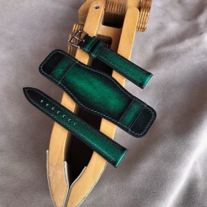 May include: Two green leather watch straps with stitching, one is a long strap and the other is a shorter strap with a buckle. The straps are displayed on a wooden surface.