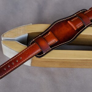 May include: A brown leather watch strap with white stitching. The strap is on a wooden tool for making watch straps.