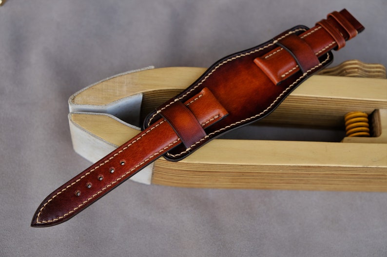 May include: A brown leather watch strap with white stitching. The strap is on a wooden tool for making watch straps.