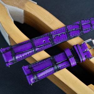 Puede incluir: Una correa de reloj de cuero morado con un patr&oacute;n de cocodrilo negro. La correa se muestra en un soporte de correa de reloj de madera.
