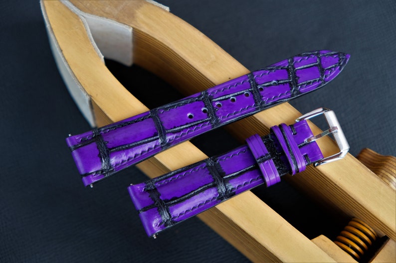 Puede incluir: Una correa de reloj de cuero morado con un patr&oacute;n de cocodrilo negro. La correa se muestra en un soporte de correa de reloj de madera.