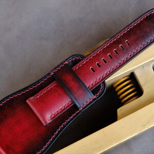 May include: A red and black leather watch strap with stitching around the edges. The strap is on a wooden tool with a spring.