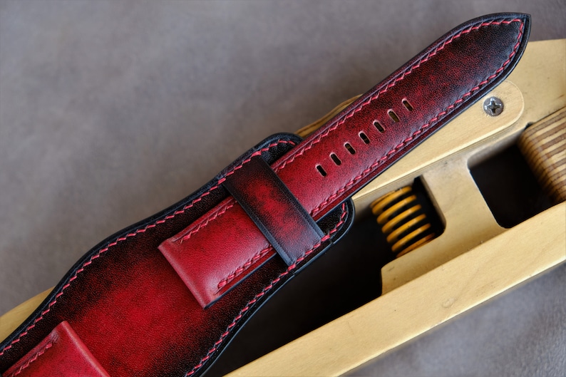 May include: A red and black leather watch strap with stitching around the edges. The strap is on a wooden tool with a spring.