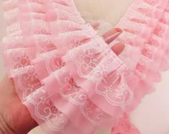 May include: Pink lace trim with multiple layers of ruffles. The lace has a delicate floral pattern.