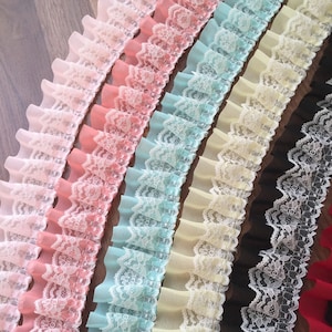 Könnte beinhalten: Eine Sammlung von fünf verschiedenen Farben von Spitzenborten mit gekräuselten Kanten. Die Spitzenborte ist weiß mit einem zarten Blumenmuster. Die Farben sind rosa, pfirsich, hellblau, creme und schwarz.