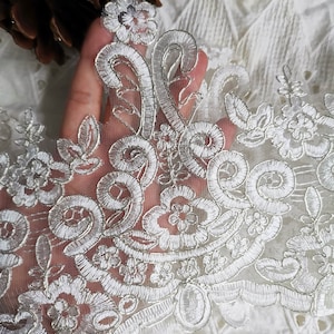 May include: Close-up of white lace trim with floral and scrollwork embroidery. The delicate design features detailed patterns and textures, perfect for crafting or embellishments. The lace is set against a soft, neutral background.