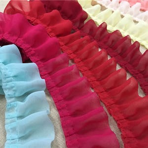 Puede incluir: Primer plano de varias filas de tela con volantes en varios colores. Los colores incluyen tonos de rojo, rosa y azul claro. La tela parece ser gasa ligera o un material similar, con una textura delicada y ondulada.