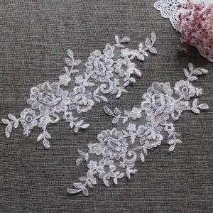 以下が含まれることがあります： 白のフローラルレースアップリケ2つ。花と葉の繊細なデザインが特徴です。グレーのテクスチャード加工された表面に配置されています。レースは複雑な模様で、衣類や工芸品の装飾に最適です。