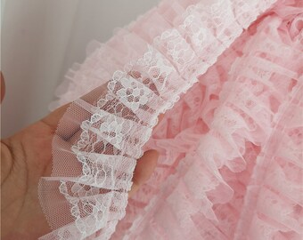 May include: White lace trim with a pink ruffled mesh fabric. The lace trim has a delicate floral pattern.