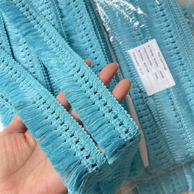 May include: Close-up of a roll of turquoise fringe trim. The trim features a woven design with a fringe edge. The trim is made of a soft, textured material. The label reads "Poly Lace Fringe".
