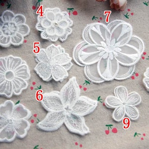 May include: Nine white lace flower appliques in diverse designs. The appliques vary in size and shape, including round, star-shaped, and multi-petal designs. The delicate lace details are visible, perfect for craft projects.