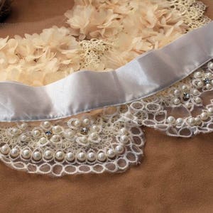 May include: White lace collar with pearl and rhinestone embellishments. The collar is attached to a white satin ribbon.