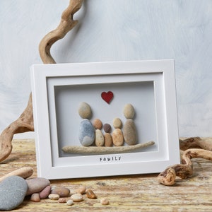 Op de afbeelding: Een wit ingelijst beeld met een familie van vijf gemaakt van kiezelsteentjes. De familie zit op een stuk drijfhout met een rood hart erboven. Het woord "FAMILIE" staat onder de familie.