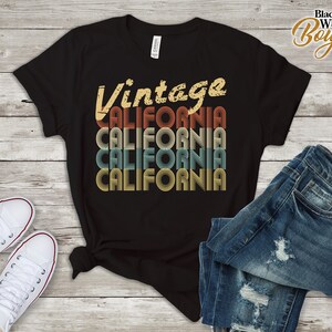 Op de afbeelding: Zwart T-shirt met een vintage-stijl grafische afbeelding met de tekst "Vintage California" in een gelaagd, retro lettertype met de kleuren rood, oranje, geel, blauw en groen.
