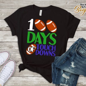Op de afbeelding: Zwart T-shirt met een wit nummer 100, groene tekst die "DAYS OF TOUCHDOWNS" zegt, en twee bruine voetballen.