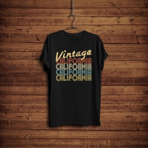 Op de afbeelding: Zwart T-shirt met een vintage-stijl grafische afbeelding met de tekst "Vintage California" in een gelaagd, retro lettertype met de kleuren rood, oranje, geel en blauw.