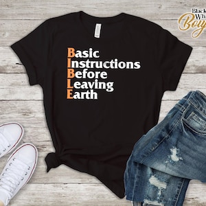 Könnte beinhalten: Schwarzes T-Shirt mit dem Akronym BIBLE in orangefarbenem und weißem Text, was für Basic Instructions Before Leaving Earth steht. Das Shirt wird mit einer blauen Jeans und weißen Turnschuhen mit roter Umrandung kombiniert.