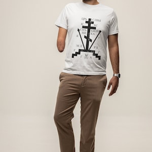 May include: A white t-shirt with a black graphic design featuring a cross with Cyrillic text. The text reads "ЦРЬ СЛВЫ ИС ХС СНЬ БЖЫЙ НИ КА К МЛ Г Г Г А РБ Г Т". The person is wearing brown pants and brown sandals.