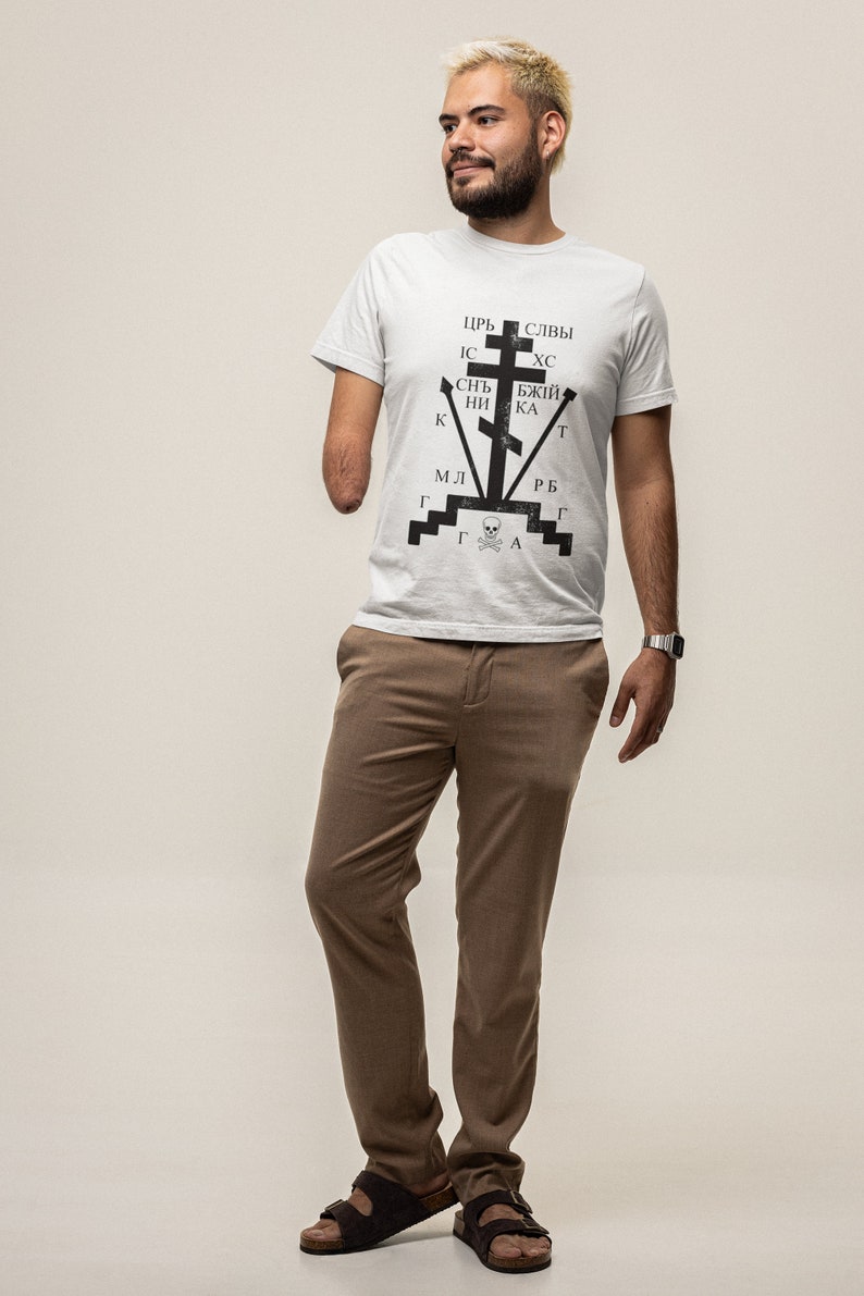 May include: A white t-shirt with a black graphic design featuring a cross with Cyrillic text. The text reads "ЦРЬ СЛВЫ ИС ХС СНЬ БЖЫЙ НИ КА К МЛ Г Г Г А РБ Г Т". The person is wearing brown pants and brown sandals.