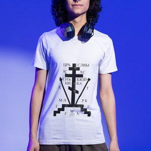 May include: A person wearing a white t-shirt with a black and white graphic of a cross and the text "ЦРЬ СЛЫВЫ ИС ХС СНТ БЖЫЙ КНИ КА Т МЛ РБ ГГГА". The person is wearing black cargo pants and headphones around their neck.