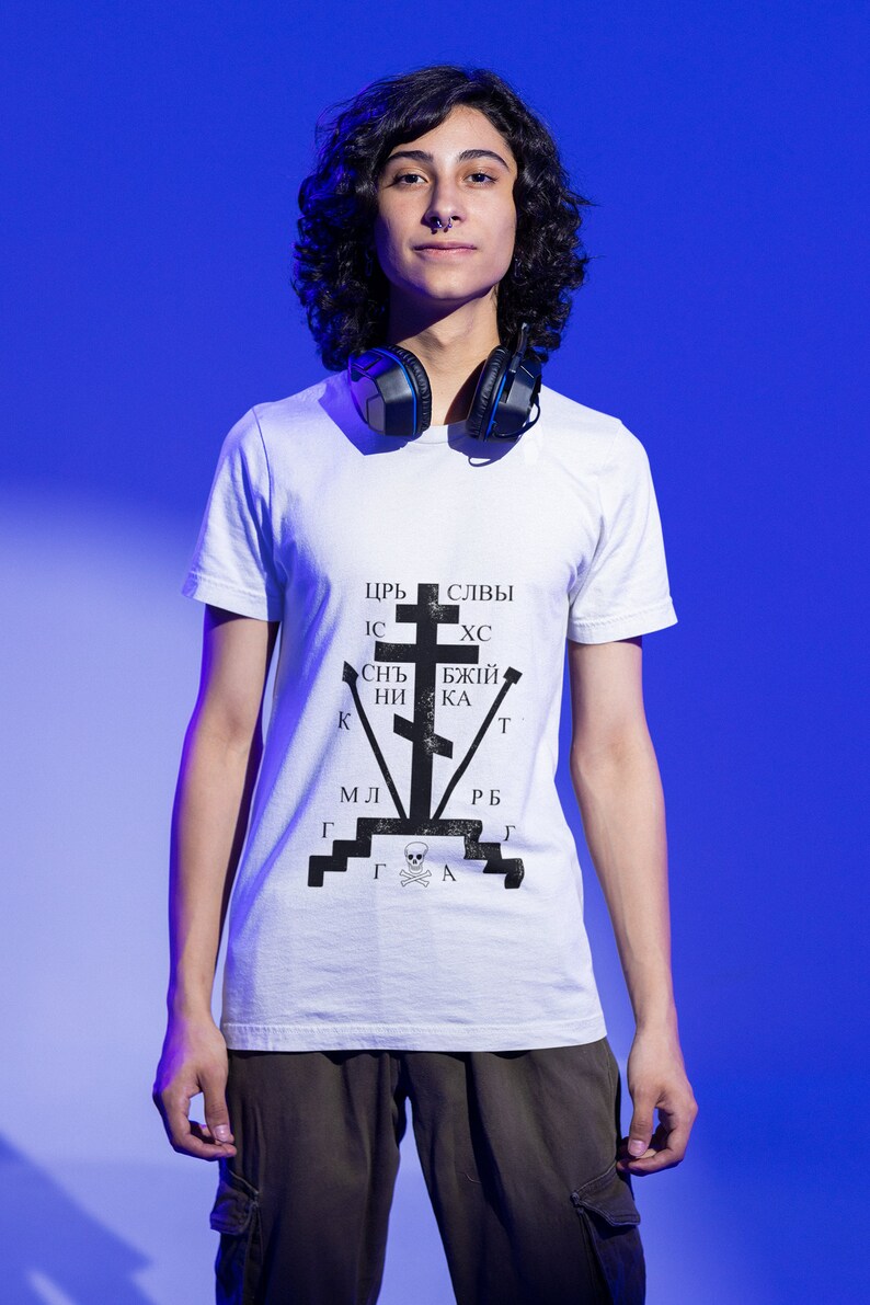 May include: A person wearing a white t-shirt with a black and white graphic of a cross and the text "ЦРЬ СЛЫВЫ ИС ХС СНТ БЖЫЙ КНИ КА Т МЛ РБ ГГГА". The person is wearing black cargo pants and headphones around their neck.