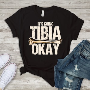 May include: Black t-shirt with a white graphic that says "It's going tibia okay" with a bone graphic. The shirt is laid out on a wooden surface with a pair of white sneakers and blue jeans.
